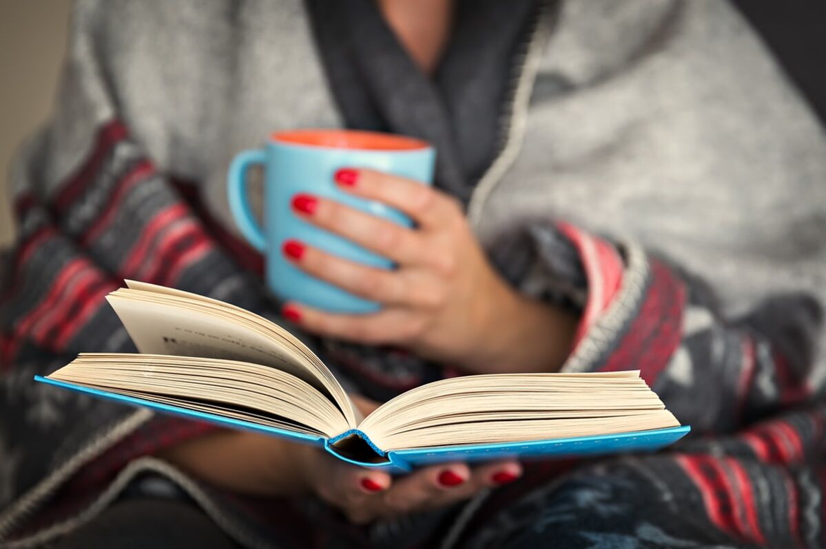 A woman reads a book and holds a hot drink